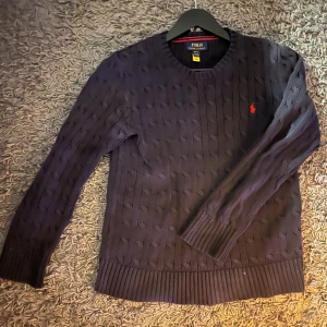 Mörkblå kabelstickad tröja från Polo Ralph Lauren - Säljer en stilren mörkblå kabelstickad tröja från Polo Ralph Lauren med det ikoniska röda logotypbroderiet på bröstet. Tröjan har långa ärmar och ribbade muddar vid ärmslut och nederkant. Perfekt för en klassisk look