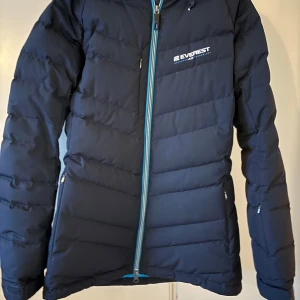 Mörkblå dunjacka från Everest - Säljer en mörkblå dunjacka från Everest med dragkedja och huva. Jackan har en stilren design med långa ärmar och är perfekt för kalla dagar. Den är utrustad med praktiska fickor med dragkedjor.