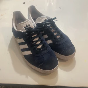 Adidas Gazelle i blå mocka - Snygga Adidas Gazelle sneakers i blå mocka med vita detaljer och snörning. De har den klassiska tre-randiga designen på sidorna och en vit sula. Perfekta för en stilren och sportig look.