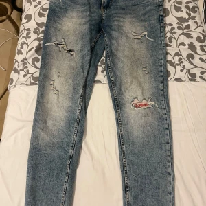 Blå slitna jeans från FSBN stolek 31/30 - Säljer ett par blå jeans från FSBN med slitna detaljer och en cool, tvättad look. De har en klassisk femficksdesign och knappgylf. Perfekta för en avslappnad stil.