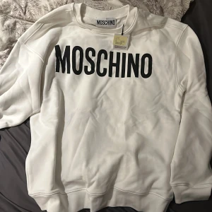 Vit sweatshirt från Moschino - Säljer en stilren vit sweatshirt från Moschino med svart logotyp på bröstet. Säljer den på grund utav att den är för liten den är storlek 50 passar även 52 helt ny med prislapp. Pris går att diskutera vid snabb affär