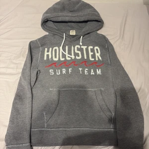 Grå hoodie från Hollister - Säljer en grå hoodie från Hollister med texten 'Surf Team' i vitt och rött. Den har en klassisk känguruficka och justerbar huva med snören. Perfekt för en avslappnad stil.