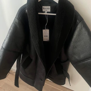 Hanna Schönberg Svart skinnjacka från NA-KD - Snygg svart skinnjacka från NA-KD med mjukt foder och bälte i midjan. Perfekt för kyligare dagar med sin stilrena design och bekväma passform. Jackan har en klassisk look med knappar framtill och två fickor.