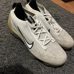 Nike Vapormax - Snygga Nike Vapormax sneakers i vitt med svart Swoosh-logga. Skorna har en stickad ovandel och genomskinlig sula med synliga luftkuddar för extra komfort. Perfekta för en sportig look.