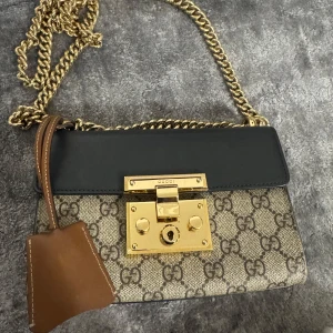 Gucci axelväska med kedja - Snygg axelväska från Gucci med klassiskt GG-mönster. Väskan har en elegant guldkedja och detaljer i svart och brunt skinn. Framsidan pryds av ett guldfärgat lås. Perfekt för att ge en lyxig touch till din outfit.