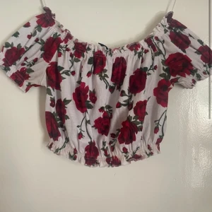 Blommig offshoulder topp från H&M Divided - Säljer en söt offshoulder topp från H&M Divided med ett blommigt mönster i rött och grönt på vit bakgrund. Toppen har korta ärmar och en elastisk kant vid axlar och midja för en bekväm passform.