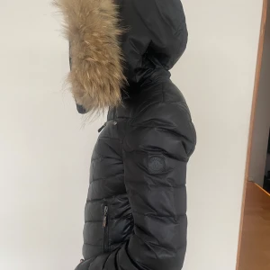 Svart Hollies pufferjacka med pälsdetaljer - Snygg svart pufferjacka med huva och pälsdetaljer. Jackan har dragkedja framtill och är perfekt för kyliga dagar. Bra vinterjacka i storlek xs med äkta pälskrage. Klotet för 3-4000