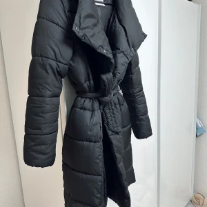 Svart lång dunjacka med bälte - Snygg och varm svart dunjacka med bälte i midjan och huva. Perfekt för kalla dagar med sin långa design och quiltade mönster. Jackan har långa ärmar och knäppning framtill. Passar perfekt för s/m/l storlek
