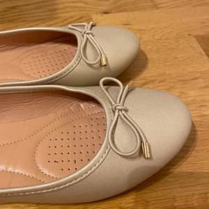 Beige ballerinaskor med rosett - Snygga beige ”guldiga” ballerinaskor med rosett. Skorna har en rund tå och en mjuk innersula för extra komfort. Perfekta för en stilren look. Jag har lagt in små kuddar vid hälarna för det blir skönare, men det går att ta bort! Jättefina men var lite för små för mig vid tårna så de är inte så använda alls!