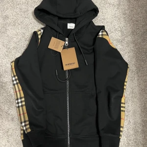 Burberry kofta - Snygg svart huvtröja från Burberry med dragkedja och klassiskt rutigt mönster på ärmarna. Tröjan har en justerbar huva och ribbade muddar. Perfekt för en stilren look.