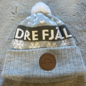 Grå mössa från Idre Fjäll - Snygg grå stickad mössa från Idre Fjäll med vit tofs och snöflingemönster. Mössan har en uppvikt kant med ett brunt märke i skinnimitation. Perfekt för kyliga dagar! Köpt i Idre vintern 2025