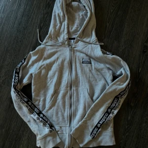 Grå hoodie från Adidas - Säljer en grå hoodie från Adidas med dragkedja och huva. Tröjan har långa ärmar med Adidas-logga längs sidorna. Perfekt för en avslappnad stil.