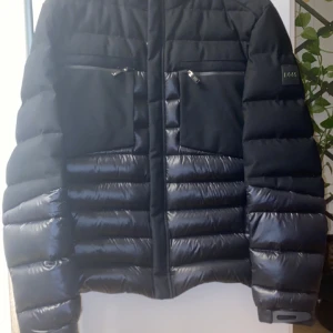 Svart pufferjacka från BOSS - Snygg svart pufferjacka från BOSS med quiltad design och dragkedja framtill. Jackan har två bröstfickor med dragkedjor och en stilren look. Perfekt för kyligare dagar. Jackan är storlek S men passar M. 