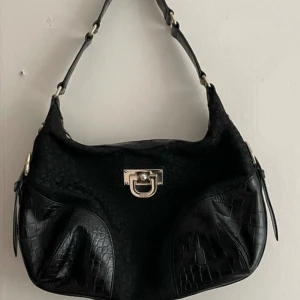 Svart axelväska med krokodilmönster - Snygg svart axelväska med krokodilmönstrade detaljer och metallspänne framtill. Väskan har en justerbar axelrem och är perfekt för att bära dina dagliga. Det finns en deformation men den är inte synlig 