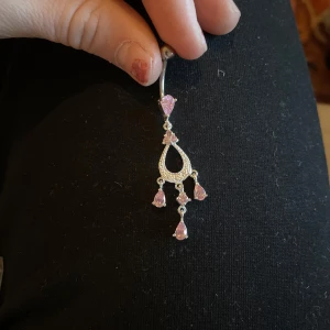 Silverfärgat navelpiercing med rosa stenar - Elegant navelpiercing i silverfärg med flera hängande rosa stenar. Designen har en droppformad detalj med gnistrande stenar som ger ett lyxigt intryck. Perfekt för att lägga till lite glamour till din stil.