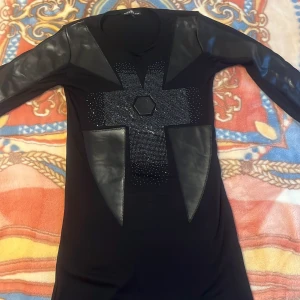 Svart klänning/tröja från Philipp Plein - Snygg svart klänning från Philipp Plein med glittrande detaljer och läderliknande inslag. Klänningen har långa ärmar och ett unikt mönster framtill med en dödskallelogga. Perfekt för en kväll ute!