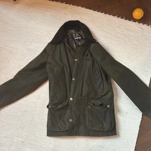 Brun  jacka från Barbour - Säljer en klassisk grön Barbour jacka med tartanmönstrat foder. Jackan har långa ärmar och stängs med både dragkedja och knappar. Perfekt för höst och vår med sina praktiska fickor och stilrena design.