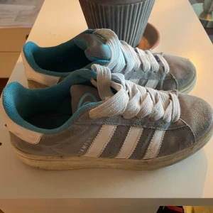 Grå Adidas Campus sneakers - Säljer ett par grå Adidas Campus sneakers med vita ränder och snörning. Skorna har en turkos insida och detaljer på tungan. Perfekta för en avslappnad stil. Skorna är i bra skick och med lite tvätt så blir dom som nya!