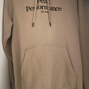 Beige hoodie från Peak Performance - Säljer en stilren beige hoodie från Peak Performance med tryck framtill. Den har en stor ficka och justerbar huva med snören. Perfekt för en avslappnad look.