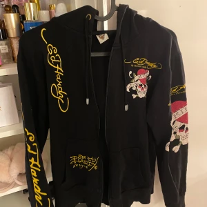 Svart hoodie från Ed Hardy - Cool svart hoodie från Ed Hardy med ikoniska tryck i gult och rött. Den har dragkedja och huva, samt detaljerade motiv av dödskallar och texten 'Love Kills Slowly'. Perfekt för en edgy look! Y2k, 