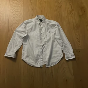 Vit skjorta från Ralph Lauren - Säljer en klassisk vit skjorta från Ralph Lauren med broderad logotyp på bröstet. Skjortan har långa ärmar och knappar framtill. Perfekt för en stilren look.
