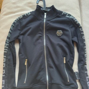 Helt ny  Philipp Plein  - Skick: 10/10 storlek: M , inga diamanter saknas!!. Legat i garderoben i flera år då jag aldrig hittar något tillfälle att ha tröjan på mig..                                                                          Snygg svart jacka från Philipp Plein med dragkedja och logotypdetaljer. Jackan har långa ärmar med mönstrade band längs sidorna och en stor logotyp på ryggen. Perfekt för en stilren look.