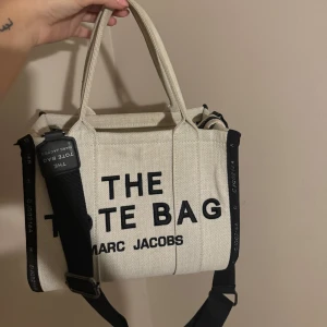 Beige tote bag från Marc Jacobs - Snygg beige tote bag från Marc Jacobs med svart text och detaljer. Väskan har både handtag och en justerbar axelrem för bekväm användning. Perfekt för att bära med sig det nödvändigaste med stil.