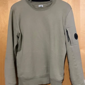 Beige tröja från C.P. Company - Snygg beige tröja från C.P. Company med rund hals och en unik ficka med dragkedja och knappdetalj på ärmen. Perfekt för en stilren och avslappnad look.