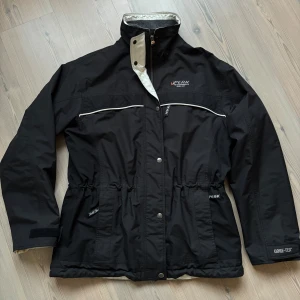 Svart jacka från Peak Performance - Säljer en svart jacka från Peak Performance i Gore-Tex-material. Jackan har dragkedja och knappar framtill, samt justerbar midja för bättre passform. Perfekt för utomhusaktiviteter med sin slitstarka design och långa ärmar.