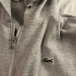 Grå hoodie från Lacoste - Säljer en klassisk grå hoodie från Lacoste med dragkedja och huva. Tröjan har det ikoniska krokodilmärket på bröstet och praktiska fickor framtill. Perfekt för en avslappnad stil. Nypris 1299kr!