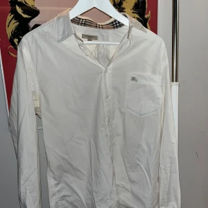 Vit skjorta från Burberry - Snygg vit skjorta från Burberry med klassiskt rutigt mönster på insidan av kragen och ärmsluten. Skjortan har en bröstficka och knappar framtill. Perfekt för en stilren look.