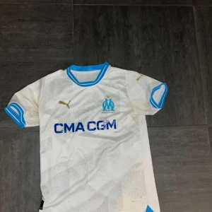 Vit fotbollströja från Puma - Säljer en vit fotbollströja från Puma med blå detaljer och klubbmärke på bröstet. Tröjan har korta ärmar och en rund halsringning med blå kant. Perfekt för fotbollsfans!