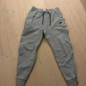 Nike tech fleece byxor - Säljer ett par grå mjukisbyxor från Nike med elastisk midja och snörning. Byxorna har en avslappnad passform och är perfekta för en chill dag. De har ribbade muddar vid bensluten och en liten Nike-logga på låret. Storlek M