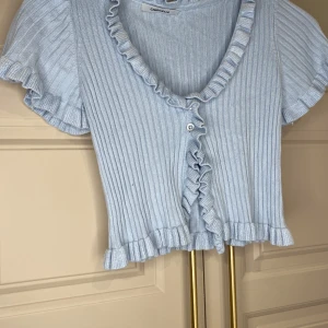 Ljusblå ribbad topp från Chiquelle - Söt ljusblå ribbad topp från Chiquelle med volangdetaljer och knappar framtill. Toppen har korta ärmar och en croppad passform, perfekt för en somrig look.