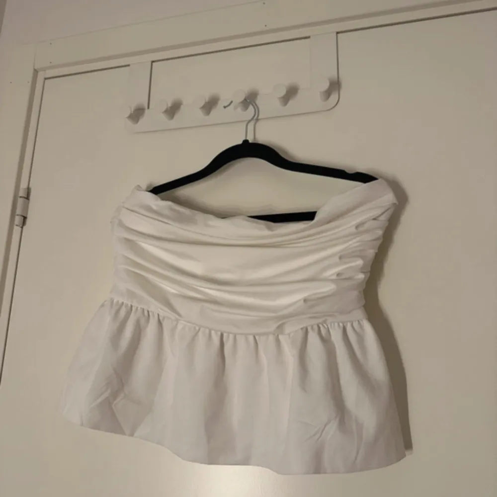 Säljer en stilren vit axelbandslös topp med volangdetalj i nederkant. Perfekt för sommarens varma dagar. Toppen har en smickrande passform och är lätt att matcha med olika outfits.. Yläosat.