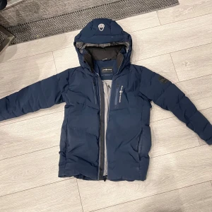 Mörkblå pufferjacka från Sail racing Antarctica - Snygg mörkblå pufferjacka från Sail racing Antarctica med huva och dragkedja. Perfekt för kalla dagar med sin vadderade design. Jackan har en logotyp på ärmen och en stilren look.