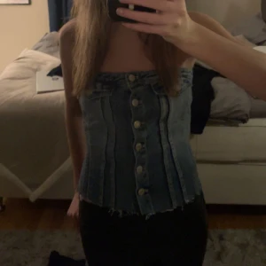 Jeanskorsett med knappar - Trendig jeanskorsett med knappar framtill. Den har en tight passform och en snygg, rå kant nedtill. Perfekt för en cool och avslappnad stil.