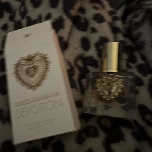 Dolce & Gabbana Devotion Eau de Parfum - Elegant parfym från Dolce & Gabbana i en lyxig flaska med gulddetaljer och ett vackert hjärtmotiv. Förpackningen är stilren med samma hjärtmotiv och märkets logotyp. En doft som utstrålar exklusivitet och passion.endast öppnad för lukt av doften som luktar. vanilj 