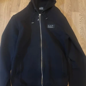 Svart hoodie från EA7 - Snygg svart hoodie från EA7 med dragkedja och justerbar huva. Perfekt för en avslappnad stil. Märkeslogga på bröstet ger en stilren touch. Passar M