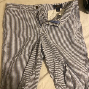 Randiga byxor från Polo Ralph Lauren - Snygga blå- och vitrandiga byxor från Polo Ralph Lauren. De har en klassisk design med dragkedja och knapp framtill. Perfekta för en stilren look till sommaren, den passar dem som har mellan s och m