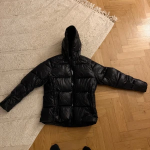 Svart pufferjacka från Save The Duck - Snygg svart pufferjacka från Save The Duck med huva och dragkedja. Jackan är 100% djurfri och har en cool logga på bröstet.  100% syntetisk dyn. 3 år gammal ungefär, använd 1 vinter sedan bara hängt i garderoben.