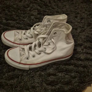 Vita Converse All Star sneakers - Säljer ett par klassiska vita Converse All Star sneakers i storlek dam 36. Skorna har en hög modell med snörning och den ikoniska röda och blå randen på sulan. Perfekta för en avslappnad stil.