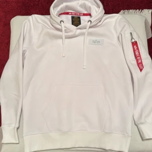 Vit hoodie från Alpha Industries - Oanvänd tröja men saknar prislappen. Säljer en stilren vit hoodie från Alpha Industries med en stor logga på ryggen och en mindre på bröstet. Hoodien har en praktisk ficka med dragkedja på ärmen och en klassisk känguruficka framtill. Den har även en röd detalj med texten 'Remove Before Flight'. Perfekt för en casual look!