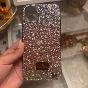 Glittrigt mobilskal från Swarovski för iPhone 11 - Snyggt och glittrigt mobilskal från Swarovski. Skalet är täckt med silverglitter och har en elegant metallplatta med Swarovski-loggan. Perfekt för att ge din telefon en lyxig touch. 