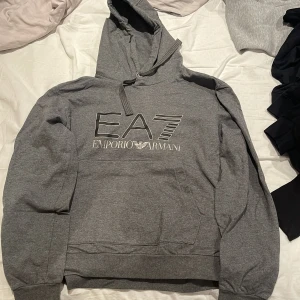 Grå hoodie från Emporio Armani - Säljer en stilren grå hoodie från Emporio Armani med EA7-logga på bröstet. Tröjan har en klassisk huva med dragsko och långa ärmar. Perfekt för en avslappnad look.