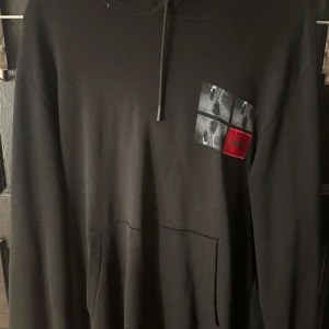 Svart hoodie från Hugo - Snygg svart hoodie från Hugo med tryck på både fram- och baksidan. Framsidan har ett fyrkantigt mönster med bilder och en röd detalj. Baksidan pryds av ett stort fototryck och texten 'HUGO' i vitt och rött. Perfekt för en cool och avslappnad stil.