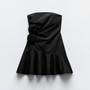 Svart axelbandslös klänning med volang - Elegant svart axelbandslös klänning med volangdetalj i nederkant. Klänningen har två dekorativa rosor på sidan som ger en unik touch. Perfekt för en stilren och sofistikerad look.