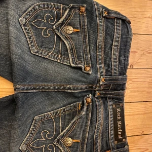 Jeans från Rock Revival - Snygga jeans från Rock Revival med unika broderade bakfickor och dekorativa knappar. De har en mörkblå tvätt och klassisk femficksdesign. Perfekta för en stilren look. Finns ett snöre till som gör så de passar om de är förstora annars är de bara att ta bort snöret. Innerbenslängden = 82 cm midjemått = 37 cm 