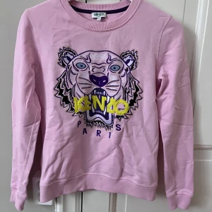 Rosa sweatshirt från Kenzo - Säljer en snygg rosa sweatshirt från Kenzo med ett coolt tigertryck i lila och gult på framsidan. Tröjan har rund hals och långa ärmar, perfekt för en avslappnad stil. Tillverkad i mjuk bomull för extra komfort.