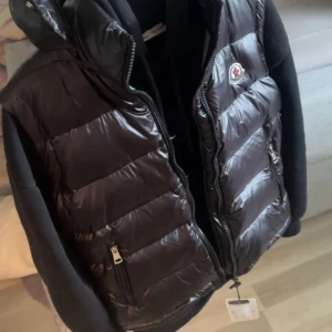 Svart dunväst från Moncler - Snygg svart dunväst från Moncler med glansig yta och dragkedja framtill. Västen har en hög krage och två praktiska fickor med dragkedjor. Perfekt för kyligare dagar.